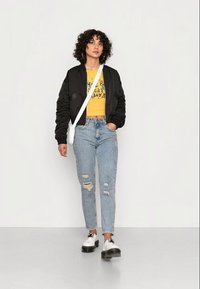 Casaco bomber preto, top cropped gráfico amarelo, jeans claros distressed, e sapatos de plataforma pretos. Bolsa de ombro simples e brincos de argola.