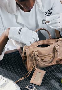 Personne portant des gants blancs examinant un sac à main en cuir beige avec une loupe sur une table à grille noire.