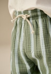 Pantalones a rayas verdes y azul claro con tela texturizada. Presentan un cordón blanco roto anudado y bolsillos laterales.