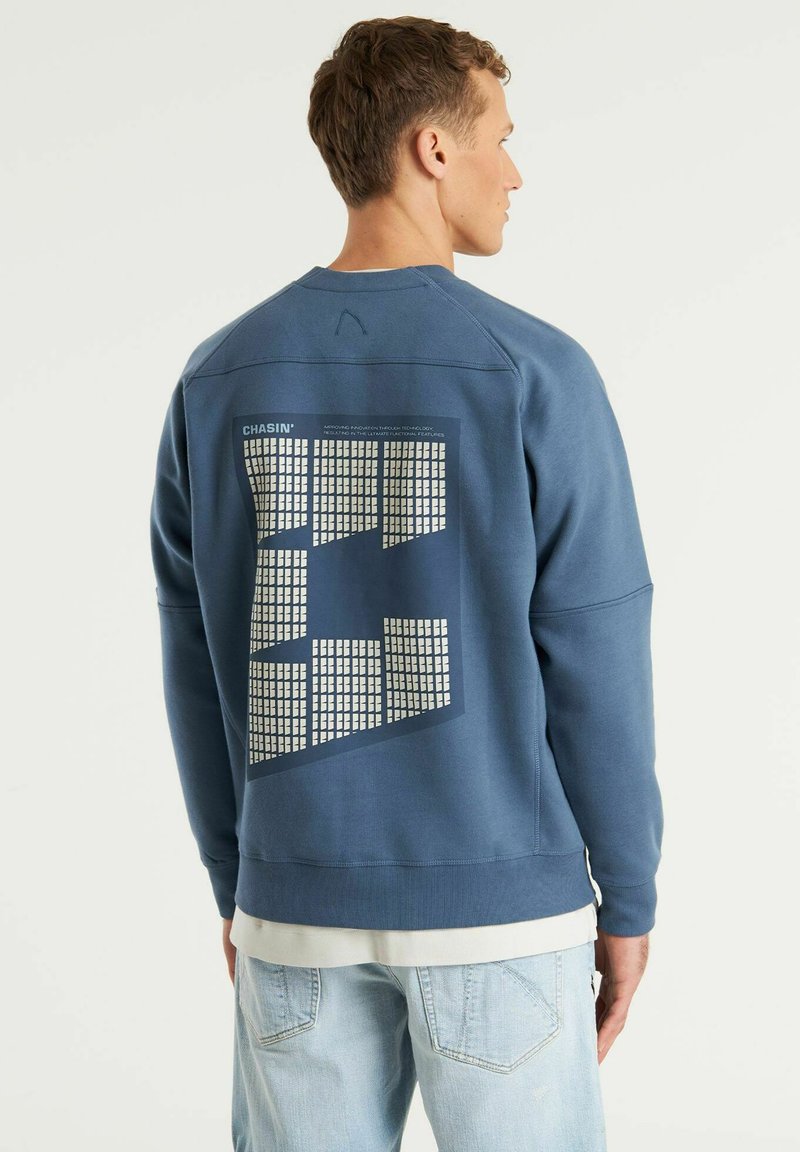 Felpa blu con tessuto testurizzato, grande design grafico sulla parte posteriore, polsini e orlo a costine, indossata sopra una maglietta di colore chiaro.