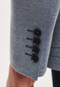 Dopasowana marynarka z rękawami w niebieskim materiale w jodełkę, z czterema czarnymi guzikami i dekoracyjnymi haftami. Gładka faktura i wyrazisty design.