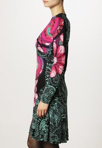 Nainen polvimittaisessa mustassa mekossa, jossa on kirkkaan vaaleanpunaisia kukkia, teal-värisiä paisleymalleja ja pitkät hihat, seisoo profiilissa valkoista taustaa vasten.