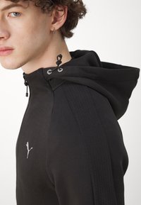 Felpa con cappuccio nera, dotata di chiusura con zip, pannelli laterali texturizzati e logo riflettente. Presenta cordini regolabili al collo.