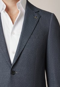 Granatowa marynarka z teksturowanej tkaniny, z czarnym guzikiem, klapami w kształcie litery V oraz małym metalowym logo po lewej stronie. Biała koszula pod spodem.