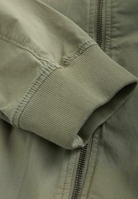 Nahaufnahme eines olivgrünen Jackenärmel mit geripptem Bündchen und sichtbarem Metallreißverschluss auf Stoffhintergrund.