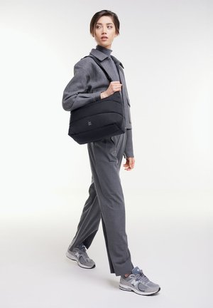 Jeune femme en tenue grise et baskets marchant, portant un sac à bandoulière noir matelassé sur un fond blanc.