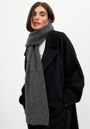 Vrouw met een lange zwarte jas en een dikke, gestructureerde donkergrijze sjaal om haar nek, tegen een effen lichte achtergrond.