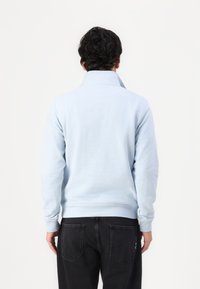 Uomo di spalle che indossa un maglione a collo alto blu chiaro e jeans neri, di fronte a uno sfondo bianco semplice.