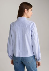 Blauw-wit gestreepte overhemd met knopen. Kenmerken zijn een kraag, een geplooide achterkant en gatherings aan de mouwen. Gemaakt van lichte stof.