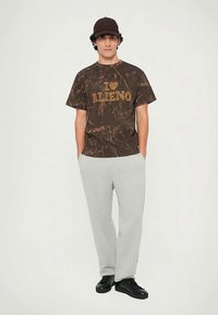 Joven con camiseta marrón con efecto tie-dye que dice "I love Milano", pantalones de chándal grises, zapatillas negras y gorra marrón, de pie con las manos en los bolsillos.