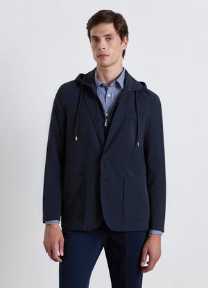 Man wearing navy blazer with hood over checked shirt standing against a plain light background.