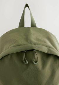 Mochila verde oliva con un diseño redondeado, que cuenta con un asa superior y un cierre de cordón. Tejido suave y texturizado con costuras reforzadas.