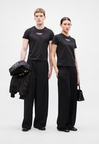 T-shirts pretas cropped com logótipo "TOMMY", combinadas com calças pretas largas. Modelo à esquerda segura uma bomber jacket preta. Estética minimalista.