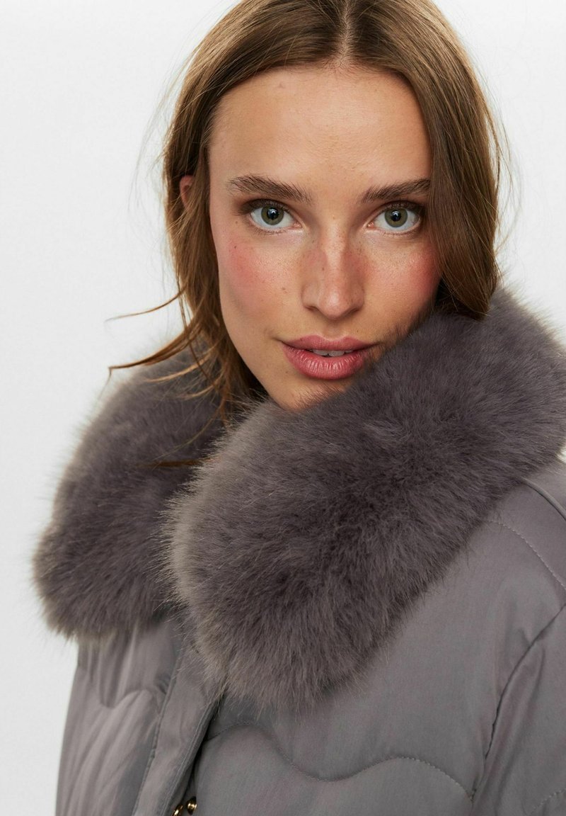 Gepolsterter grauer Mantel mit einem großen, weichen grauen Pelzkragen. Das Design umfasst eine gesteppte Textur und einen Reißverschluss mit goldenen Akzenten.