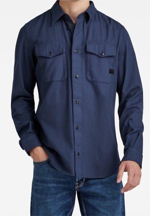 Homme portant une chemise bleu marine à boutons avec deux poches avant à rabat et un jean bleu, une main dans la poche, sur fond blanc.
