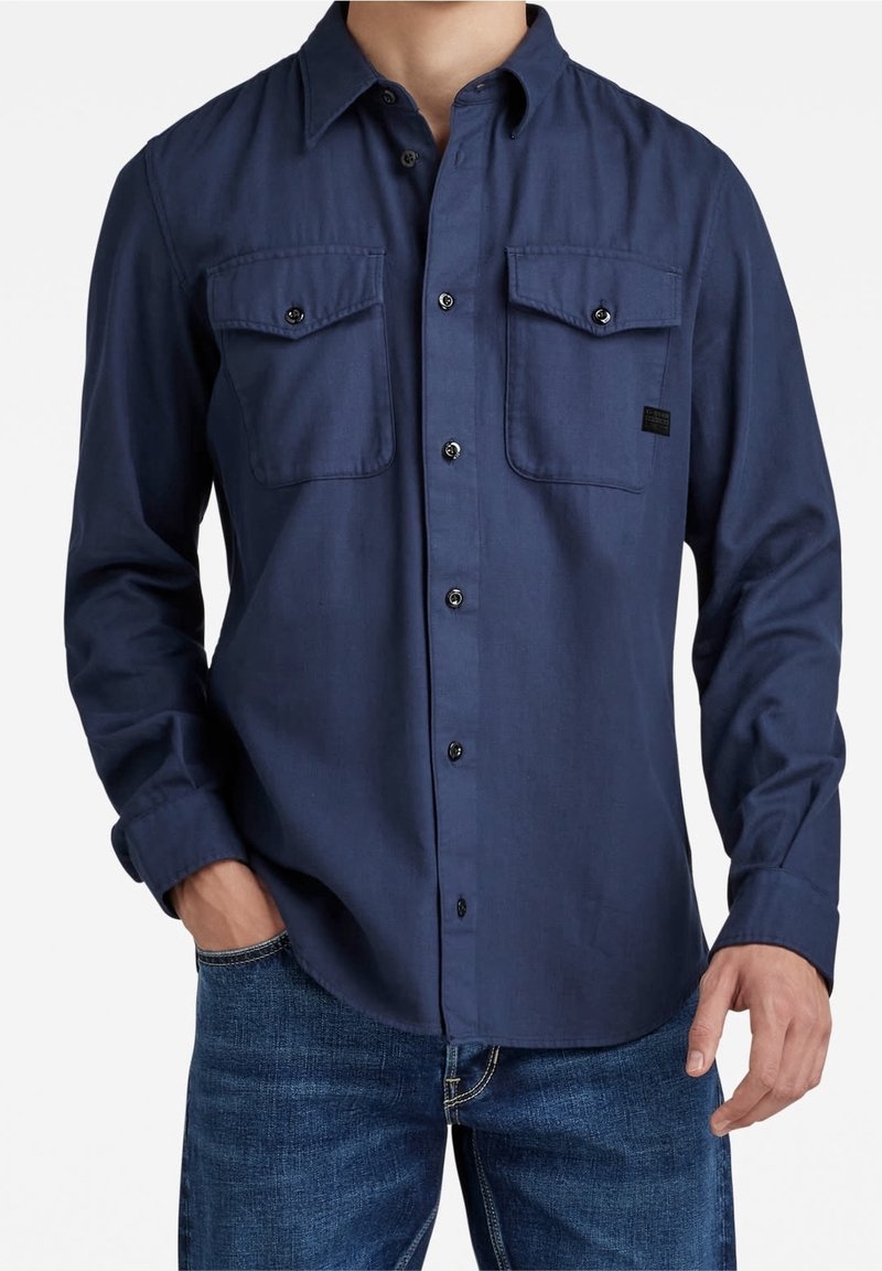 Homme portant une chemise bleu marine à boutons avec deux poches avant à rabat et un jean bleu, une main dans la poche, sur fond blanc.