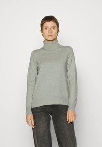 Hellgrauer Rollkragenpullover aus weichem Stoff, mit langen Ärmeln und legerem Schnitt. Kombiniert mit dunklen Jeans.