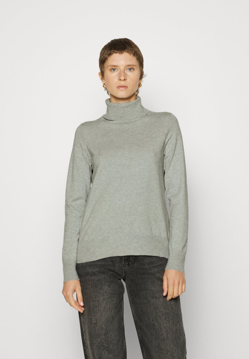 Pull à col roulé gris clair en tissu doux, avec des manches longues et une coupe décontractée. Porté avec un jean noir.