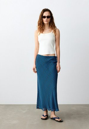 Junge Frau mit Sonnenbrille, weißem ärmellosem Oberteil, blau-schwarz wellenförmig gestreiftem langem Rock und schwarzen Flip-Flops, stehend vor einer schlichten Wand.