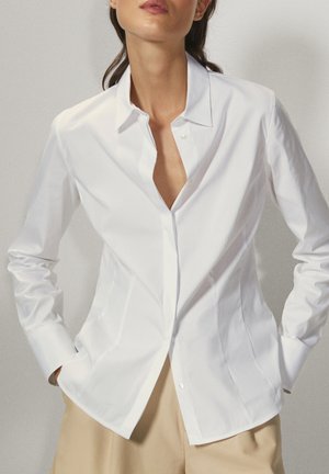 Kvinde iført en tætsiddende hvid skjorte med knapper og krave samt beige bukser, stående med hænderne i lommerne mod en ensfarvet baggrund.