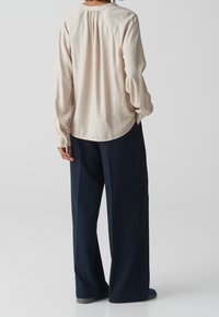 Blusa beige a maniche lunghe con orlo arrotondato e piega sul retro, abbinata a pantaloni larghi blu navy. Tessuto morbido con polsini abbottonati e vestibilità comoda.