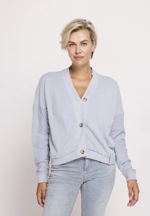 Frau mit kurzen blonden Haaren, die eine hellblaue, geknöpfte Strickjacke und hellblaue Jeans trägt, vor einem schlichten weißen Hintergrund stehend.