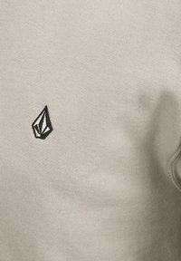 Lichtbeige sweatshirt met een gladde textuur, voorzien van een klein zwart geometrisch logo op de linkerkant van de borst. Eenvoudig, minimalistisch ontwerp.