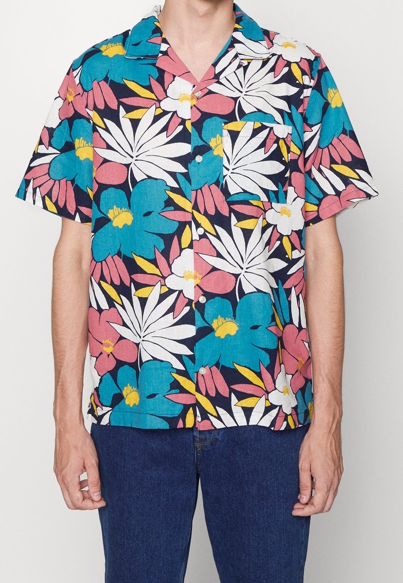 Homme portant une chemise à manches courtes boutonnée avec un motif floral vibrant bleu, blanc, rose et jaune, associée à un jean bleu foncé.