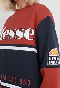 Czerwony i granatowy sweter z wyraźnym białym napisem, poziomymi paskami oraz naszywką z logo na rękawie. Materiał bawełniany, luźny krój.