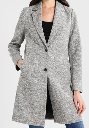 Cappotto grigio con texture, colletto a pastrano, chiusura frontale con bottoni e tasche laterali. Presenta un taglio sartoriale, raggiungendo una lunghezza fino a metà coscia.