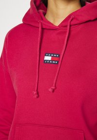 Červená mikina s kapucí vyrobená z bavlněné směsi, s přední kapsou, nastavitelnou šňůrkou a malým vyšívaným logem Tommy Jeans na hrudi.