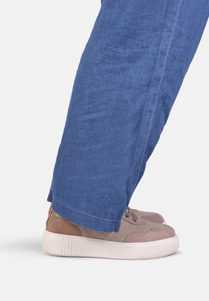 Personne portant un pantalon ample en denim bleu et des baskets en daim taupe avec des semelles blanches épaisses, debout sur un fond blanc.