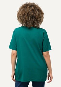 T-shirt verde in cotone con maniche corte, retro leggermente allungato, collo a giro e dettagli delle cuciture visibili. Indossato con jeans blu.