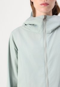 Casaco leve de cor azul claro, com capuz, detalhe de fecho em metal prateado e textura suave. Projetado para uma silhueta ajustada.