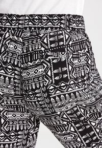 Pantalon à motifs noirs et blancs avec un design tribal, doté d'une ceinture élastique et d'une poche arrière. La texture semble lisse.