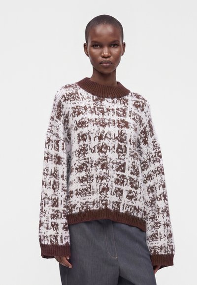 Personne aux cheveux courts portant un pull en maille marron et blanche à motif, avec des poignets et un bas côtelés, associé à un pantalon foncé.