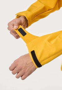 Kädet säätävät mustia tarranauhoja keltaisen takin hihassa valkoista taustaa vasten.