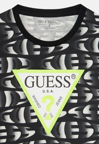 T-shirt en coton noir avec des motifs de logo blancs et gris, présentant une étiquette triangulaire audacieuse en néon vert, détaillant les informations sur la marque et le produit.