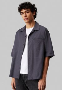 Grijze korte mouwen shirt van een lichtgewicht stof, met een relaxte pasvorm, een borstzak en zwarte knopen. Draagt over een wit t-shirt.