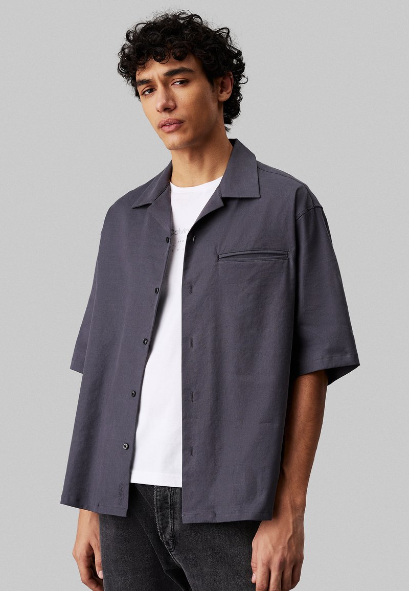 Grijze korte mouwen shirt van een lichtgewicht stof, met een relaxte pasvorm, een borstzak en zwarte knopen. Draagt over een wit t-shirt.