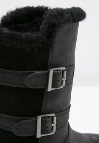 Botte en daim noir avec une doublure en fourrure noire douce, dotée de deux boucles argentées ajustables sur le côté pour la fermeture et l'ajustement.