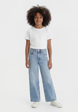 Dieťa kučeravých vlasov, oblečené v bielom tričku s krátkym rukávom, svetlomodrých širokých džínsach a bielych teniskách, stojí pred jednofarebným pozadím.