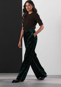 Femeie care merge îmbrăcată cu un top scurt de culoare maro închis, pantaloni cu talie înaltă din catifea verde, largi, și mule cu toc de culoare albastru marin, decorate cu fundițe.