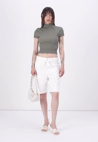 Olivengrønn, ribbestrikket, kort topp, hvite skreddersydde shorts, holder en liten veske, iført lette sandaletter. Rene linjer og moderne design.