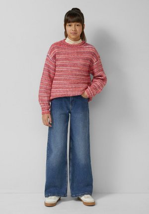 Czerwony i biały sweter z wzorem, z opuszczonymi ramionami, zestawiony z szerokimi niebieskimi dżinsami. Modelka stoi swobodnie, z jedną ręką na biodrze.