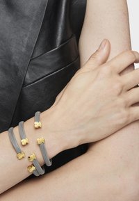 Tres pulseras de malla gris adornadas con adornos dorados en forma de pequeños ositos de peluche descansan en la muñeca de un modelo, sobre un fondo de cuero negro.