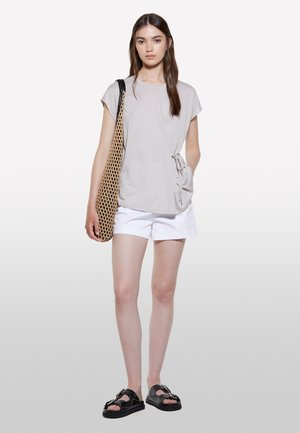 Light grey short-sleeve top with a side tie detail, paired with white shorts and black sandals. The model carries a woven tan bag.