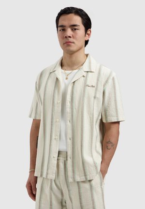 Junger Mann trägt cremefarbenes kurzärmeliges Hemd mit Knöpfen und passende Hose mit vertikalen grünen und braunen Streifen, steht mit einer Hand in der Tasche.