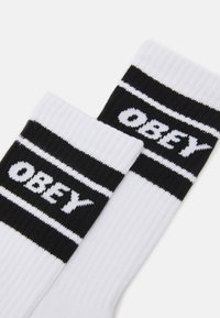White socks with black ribbed cuffs featuring horizontal black stripes and the word "OBEY" in bold white text on each sock.