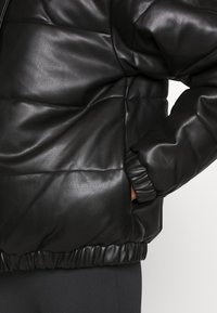 Veste en cuir noir matelassée avec un design en piquage, des poignets élastiqués et un ourlet. Texture lisse avec une finition légèrement brillante.